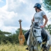 girl on a grey horse standing and looking at a giraffe behind her