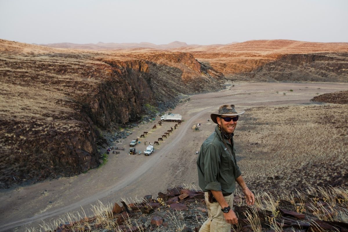 man standing on top of a rocky hill looking down over the horses eating at camp below