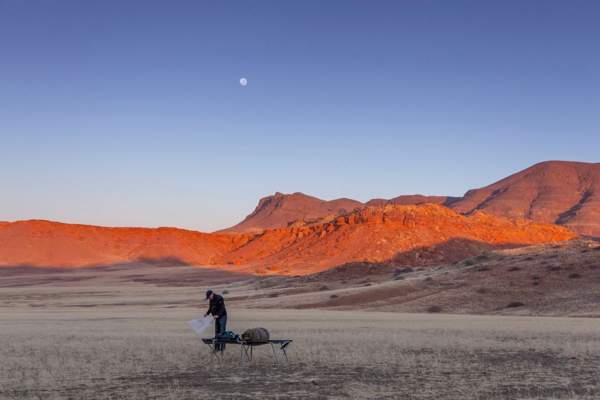 man setting up camp in damaraland, namibia below the moon with the rocky mountains behind him