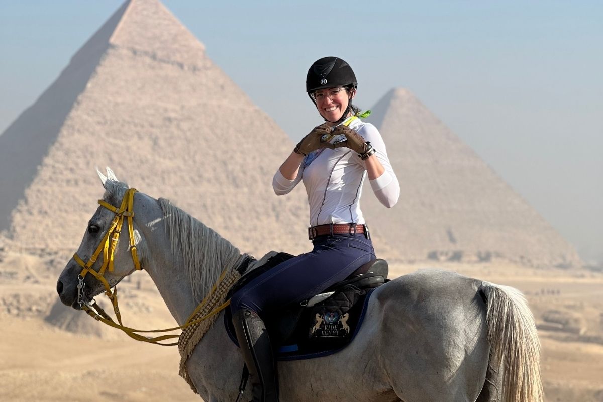 girl on grey arab horse showing heart sign with her hands with pyramids in the background in egypt