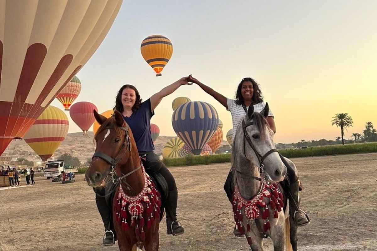 riders on horses holding hands and smiling with hot air balloons in the background at sunrise in egypt